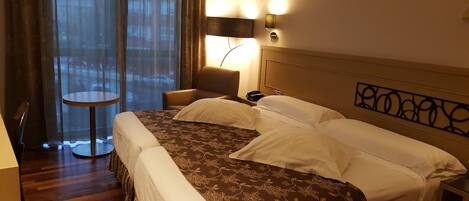 A hotel room with a large bed, a round table, a chair, and a view through sheer curtains.