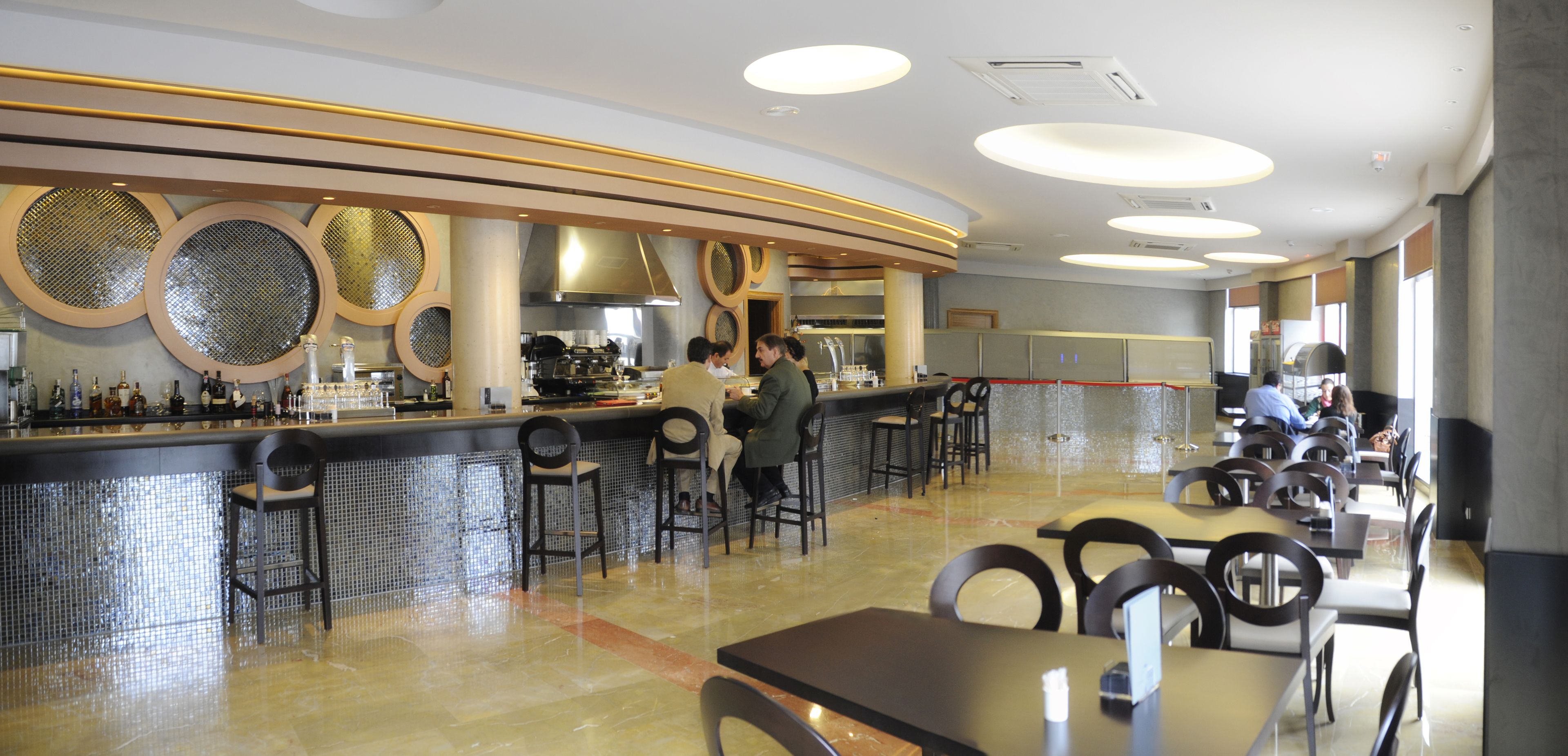 Un restaurante moderno con barra, diferentes disposiciones de asientos y una cocina visible al fondo.