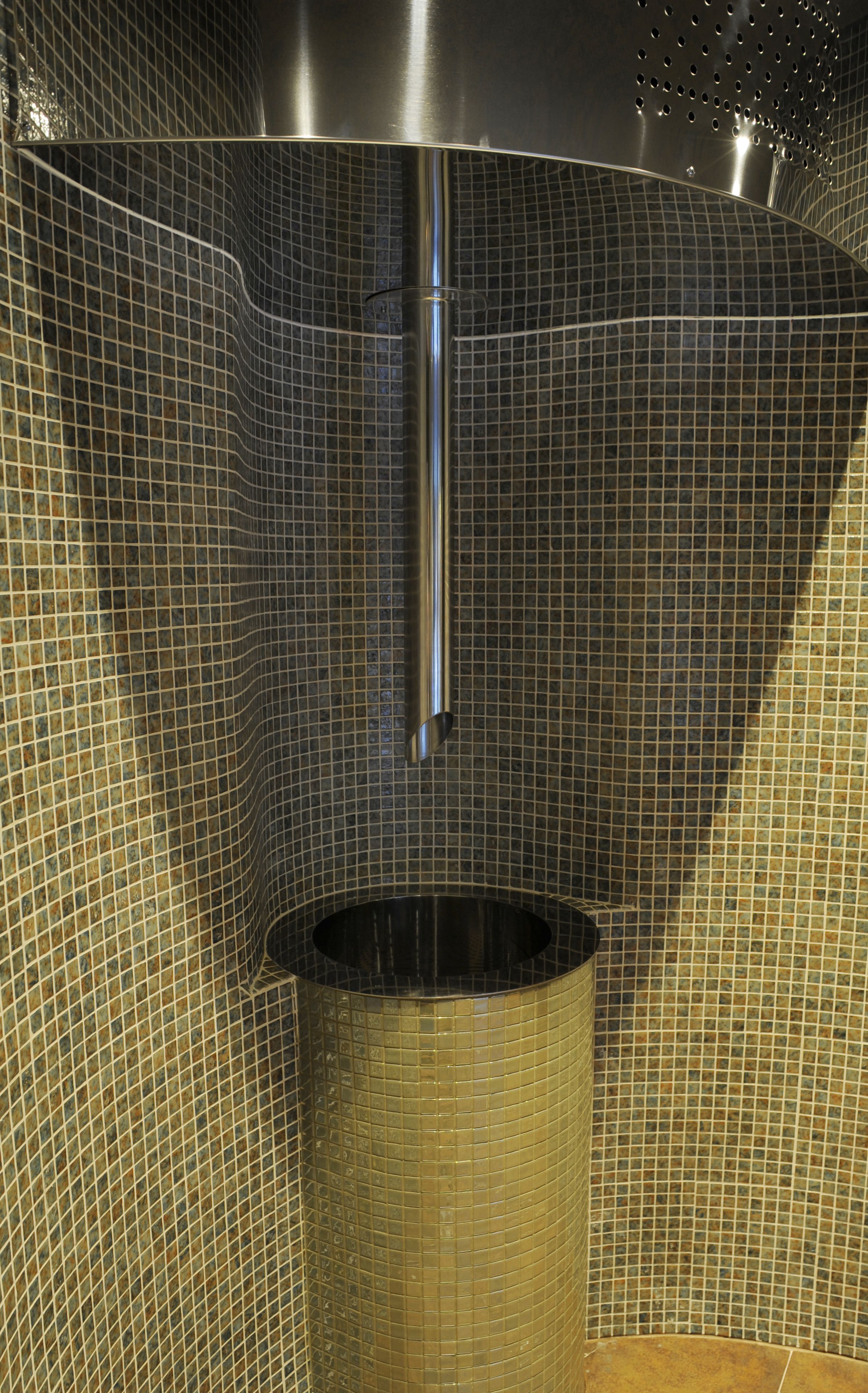 A bathroom with a tiled wall, a metallic shower head, and a cylindrical sink.