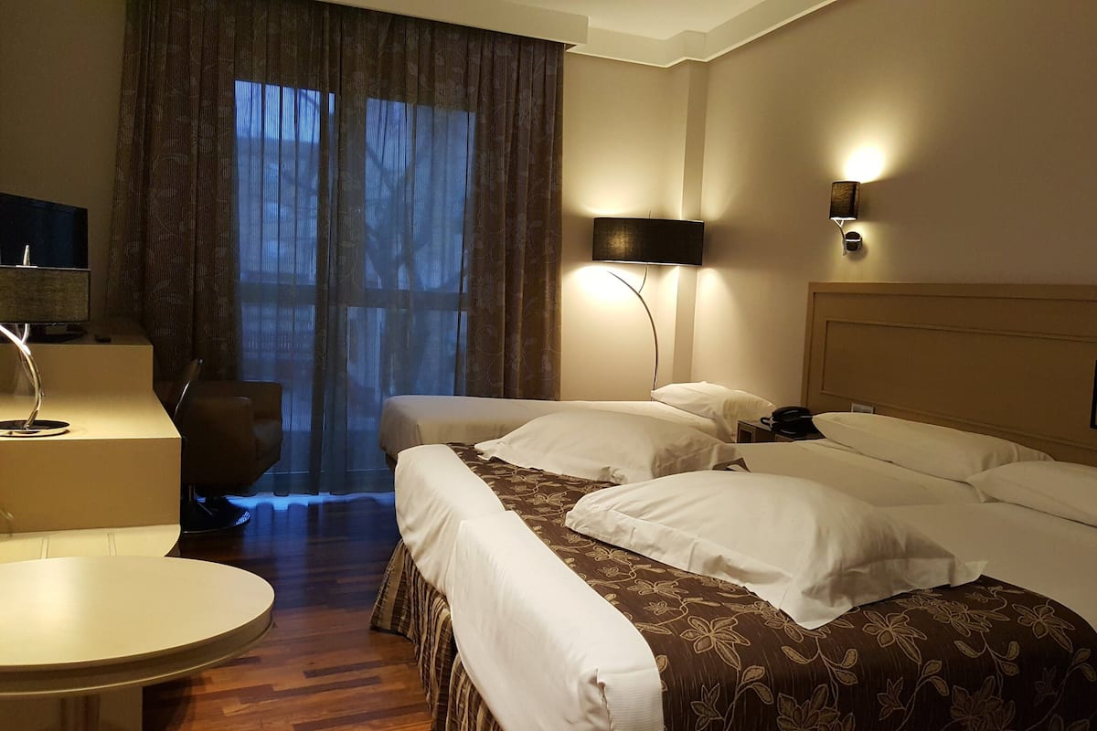 A hotel room with two beds, a desk, a chair, a lamp, and a large window with curtains.
