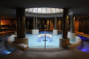 Una piscina interior moderna con una zona de estar circular central, rodeada de altos pilares y un tragaluz superior.