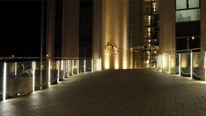 A nighttime view of a hotel entrance with a glass facade, a central logo, and a curved pathway leading to the entrance. - Cumbria Spa & Hotel (Ciudad Real)