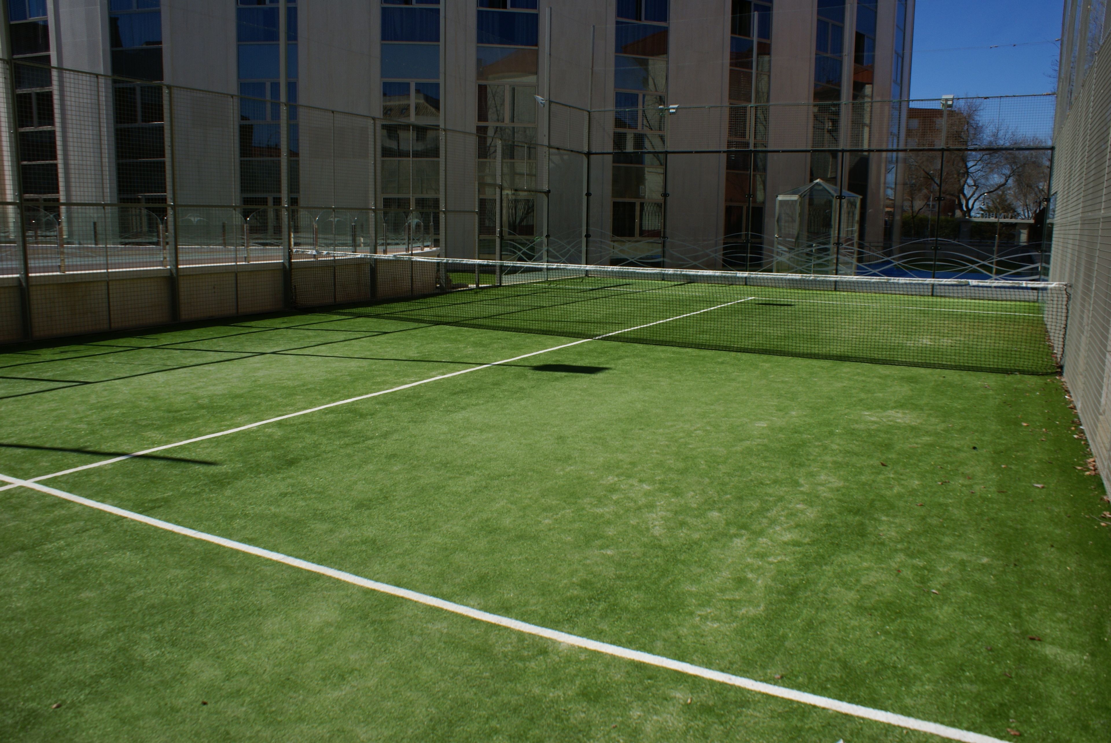 Una cancha de tenis interior con red, rodeada de paredes de vidrio y un edificio al fondo.