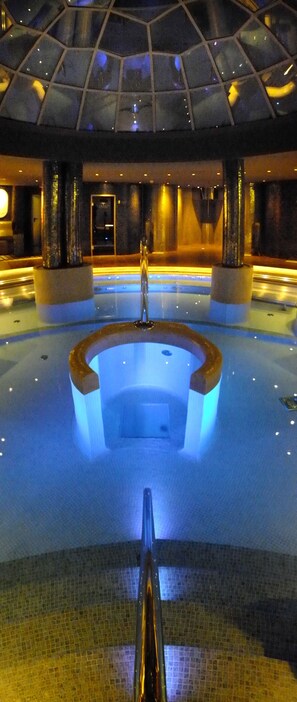 Una amplia piscina interior con una estructura circular central, rodeada por un suelo de baldosas y un techo de cristal.