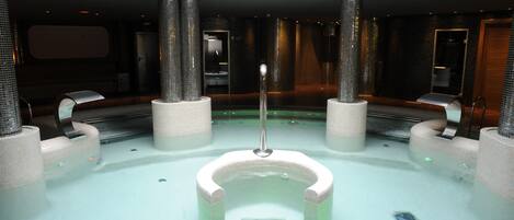 Piscina interior moderna com uma fonte central, rodeada por áreas de estar e teto alto.