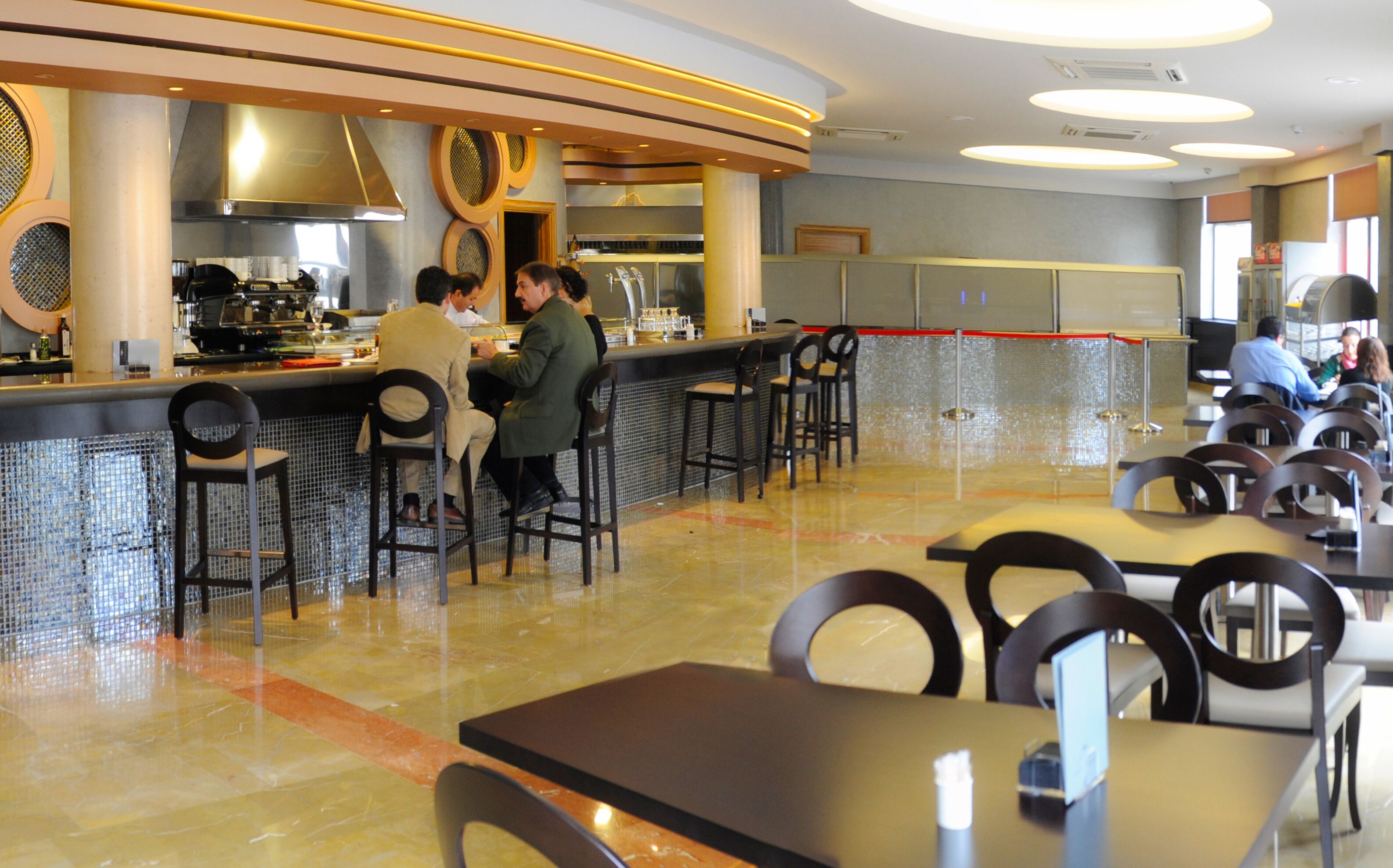 A modern restaurant with a bar area, seating arrangements, and a kitchen in the background.
