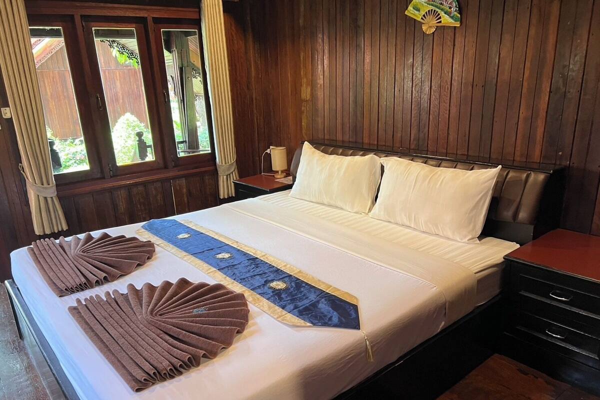 Una cama bien tendida con un corredor decorativo azul y dorado, dos toallas marrones y almohadas blancas en una habitación con paneles de madera y una ventana con cortinas.