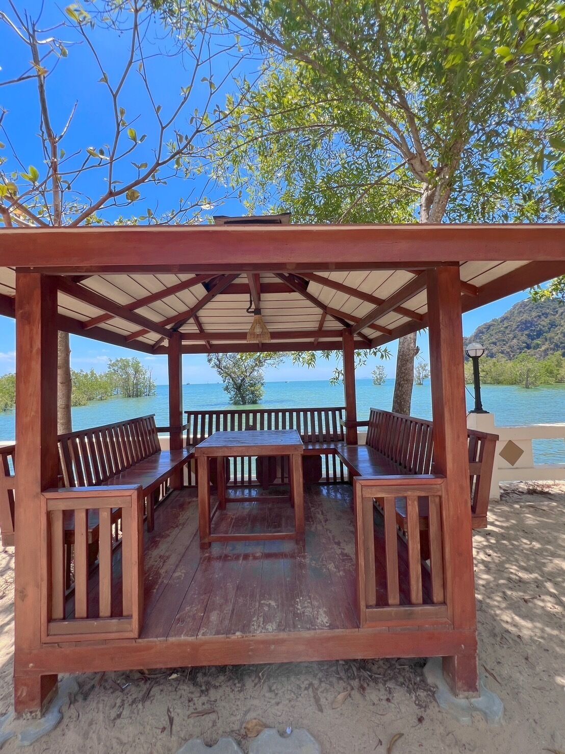 A wooden gazebo with benches and a table, overlooking a body of water and a mountainous landscape.