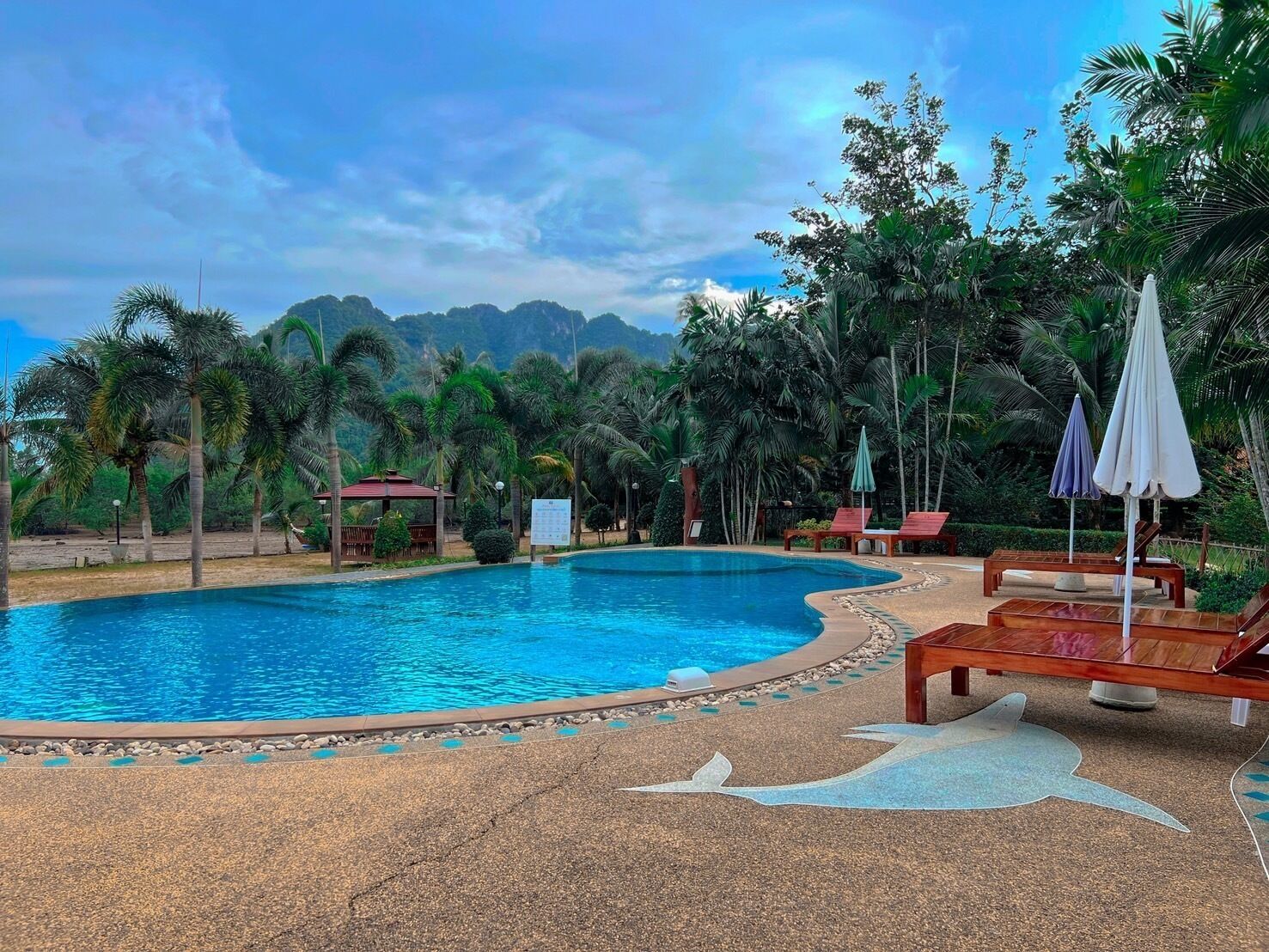 Una piscina con bordo in pietra, circondata da lettini e ombrelloni, con palme e montagne sullo sfondo.