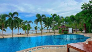Una piscina con cubierta de madera y sillas de descanso, rodeada de palmeras y vista a la playa.