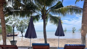 Un salón junto a la playa con sillones de madera y taburetes de plástico blanco, cubierto por sombrillas oscuras, con vista a un mar tranquilo.