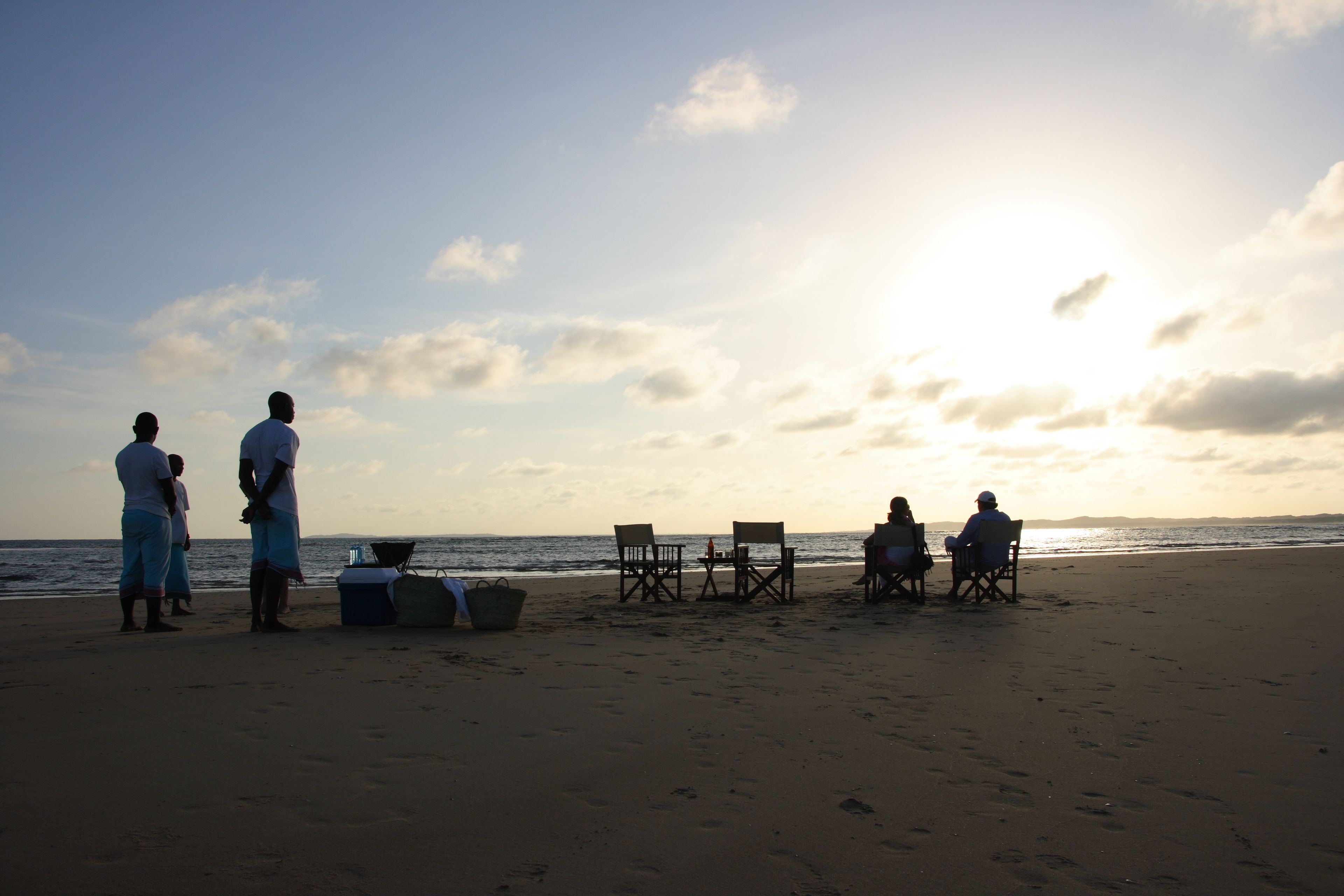 A beach scene with people standing and sitting on chairs, a table, and a cooler on the sand.