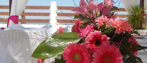 Una mesa con un arreglo floral rosa, mantel blanco y una estructura de pérgola de madera al fondo.