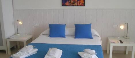 Habitación de hotel con cama, almohadas azules, cabecera blanca, dos mesitas de noche con lámparas y una obra de arte colorida en la pared.