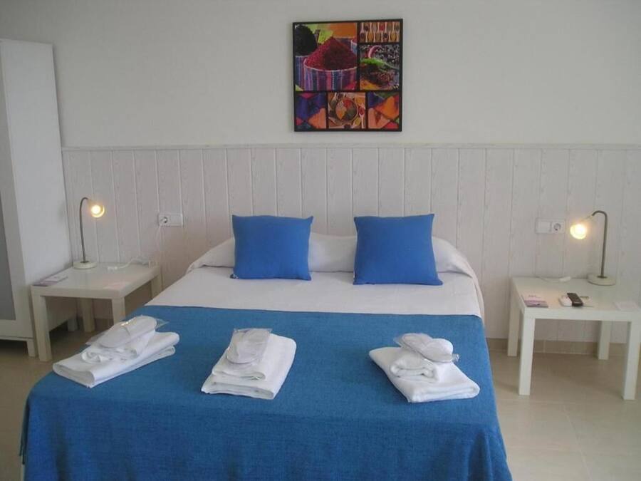A hotel room with a bed, blue pillows, white headboard, two bedside tables with lamps, and a colorful artwork on the wall.