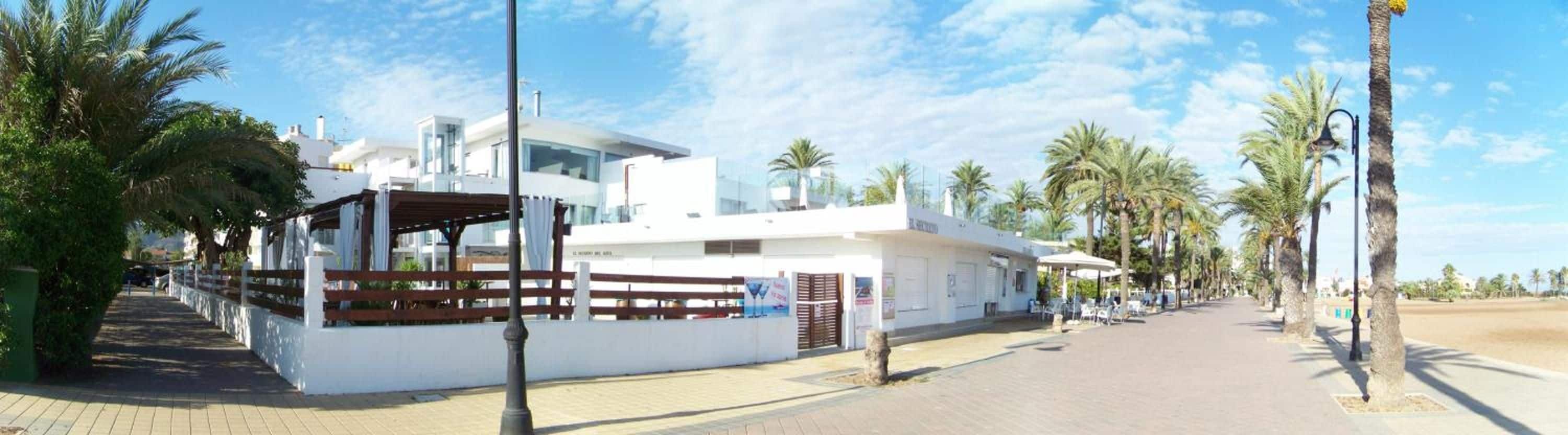 A modern building with a covered outdoor seating area, palm trees, and a clear sky.