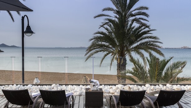 A deck with four lounge chairs, a small table, and a bottle of champagne. The deck overlooks a beach with white pebbles and palm trees. - El Secreto del Agua (Cartagena)