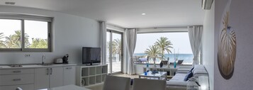 Una habitación de hotel moderna con cocina, televisor, sofá y balcón con vistas al mar y palmeras.