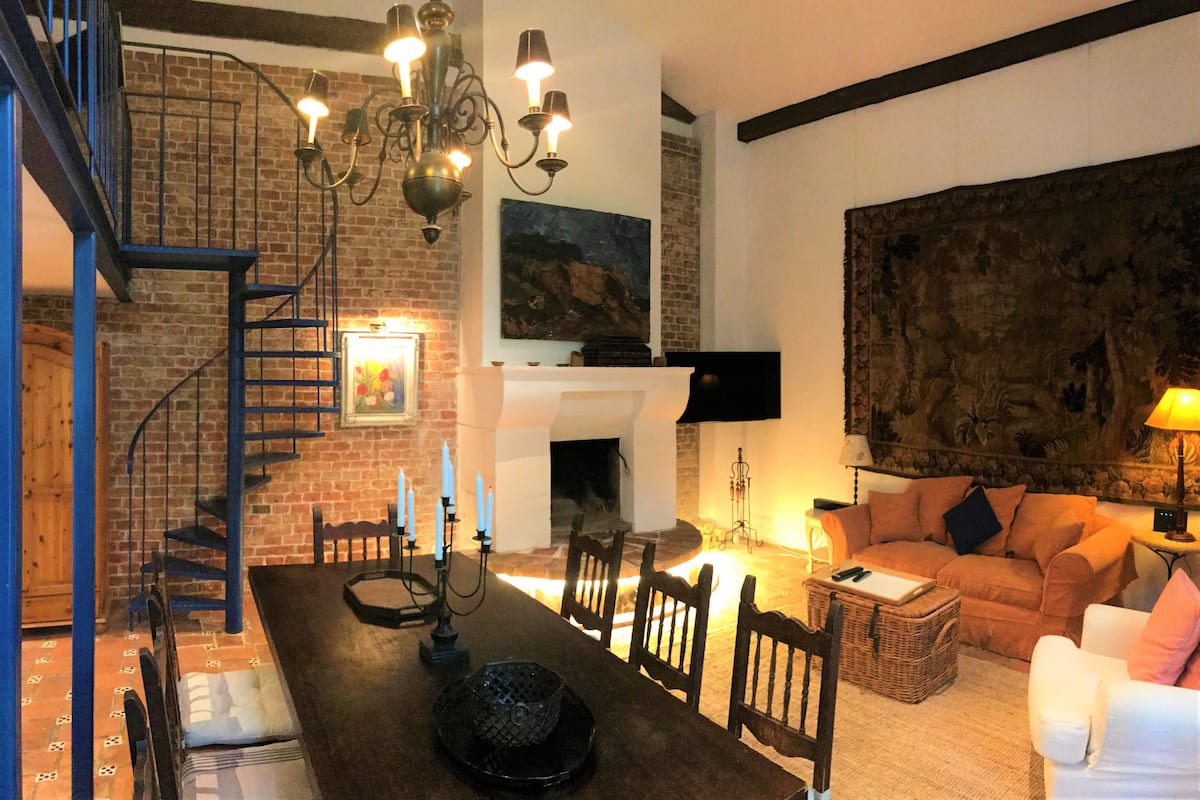 A cozy living room with a fireplace, a dining area with a long table, and a staircase leading to an upper level.
