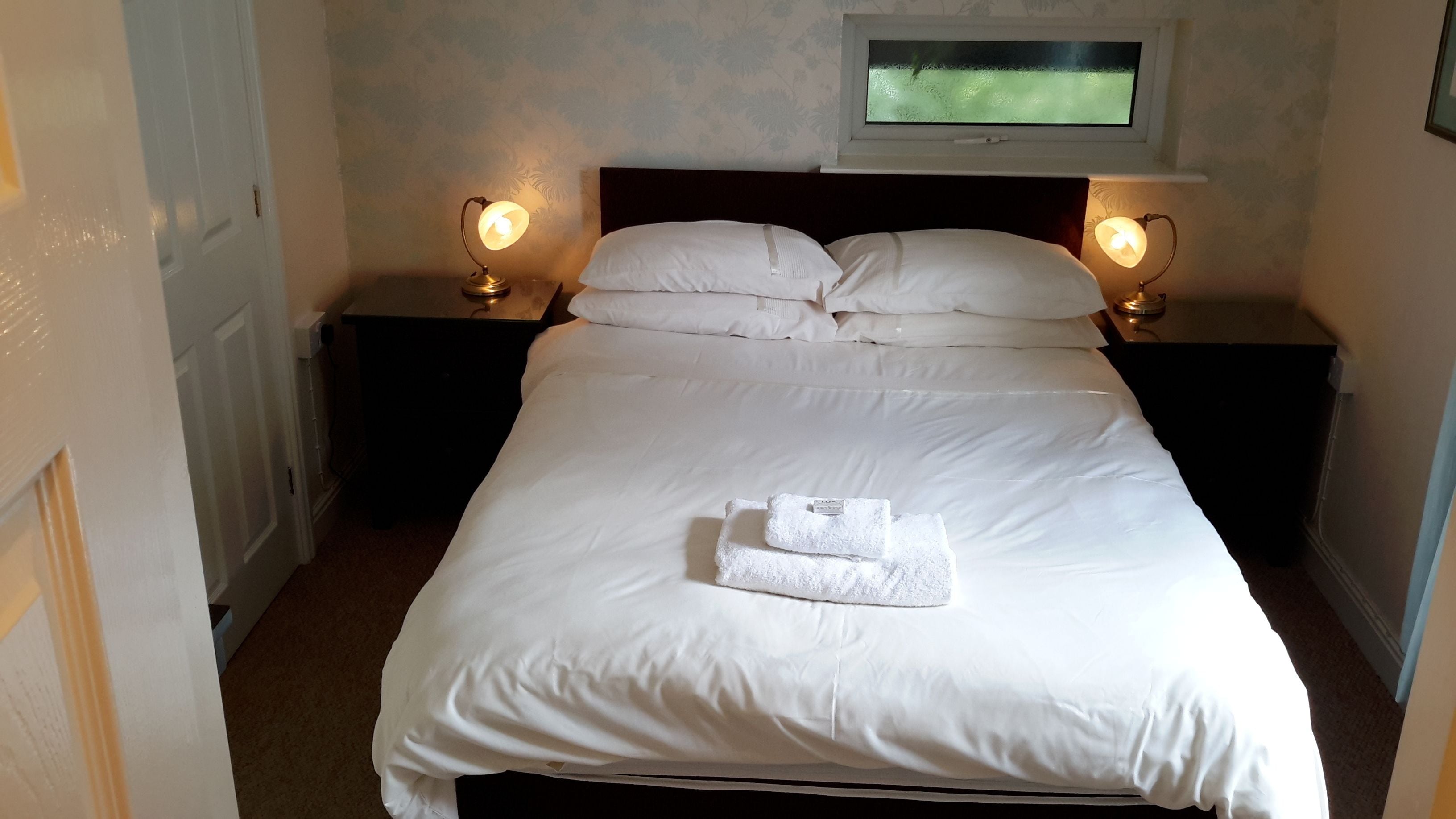 A neatly made bed with white linens and a folded towel, flanked by two bedside lamps on dark nightstands.