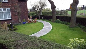 Un jardín con un sendero de piedra curvado, una mesa y sillas de madera, y una casa de ladrillo.