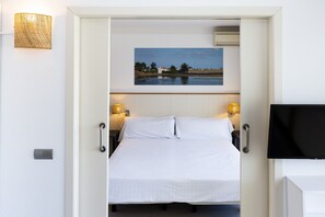 A hotel room with a bed, two bedside lamps, a flat-screen TV, and a painting of a beach scene on the wall. - Acacias Apartamentos Salou (Salou)