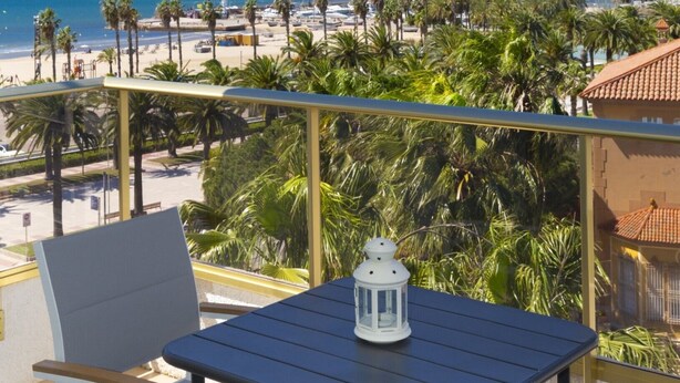 Un balcón con mesa y sillas, con vista a una playa con palmeras y un cielo azul despejado.