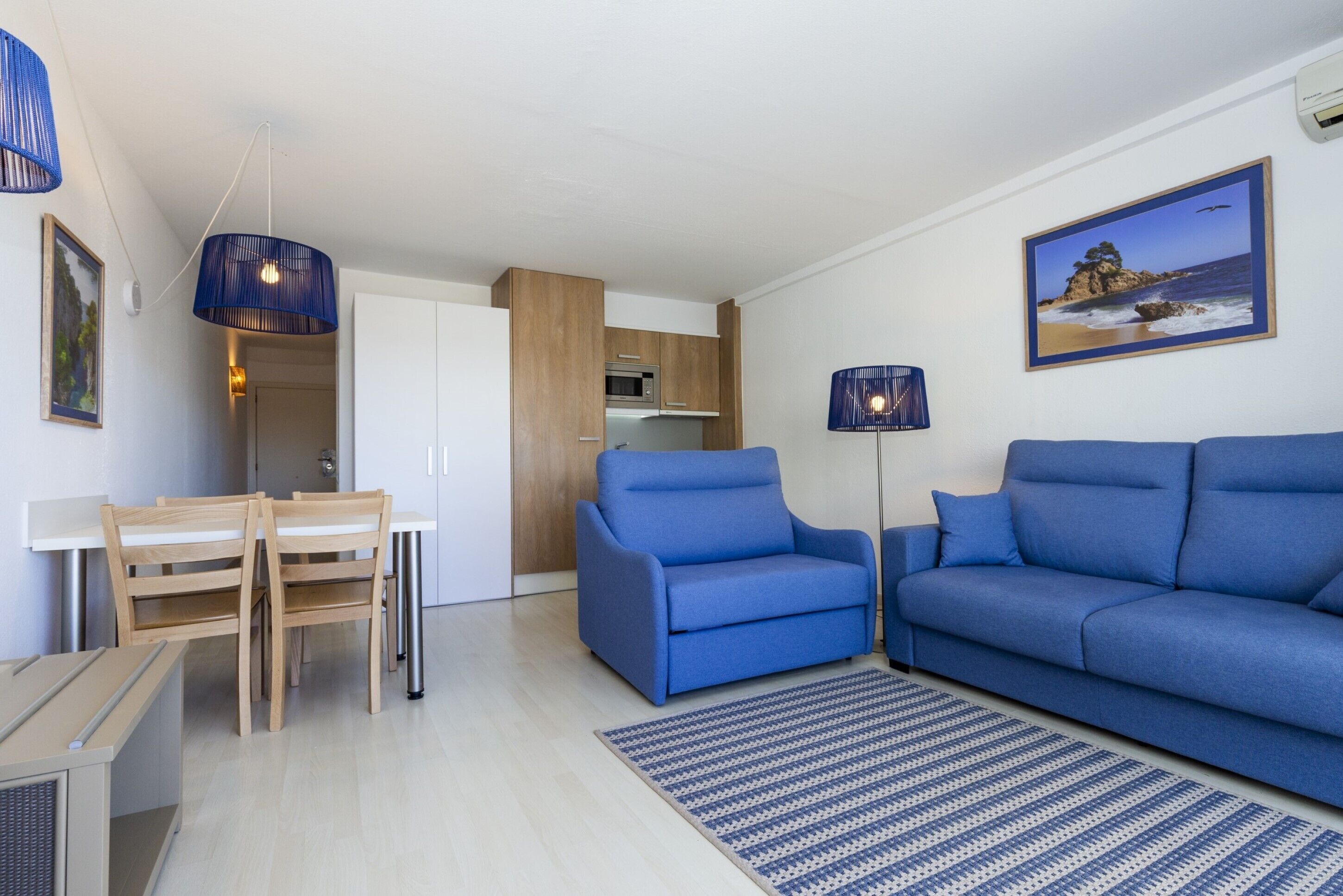 A modern living room with a blue sofa, a dining table with chairs, a kitchen area, and a framed picture on the wall.