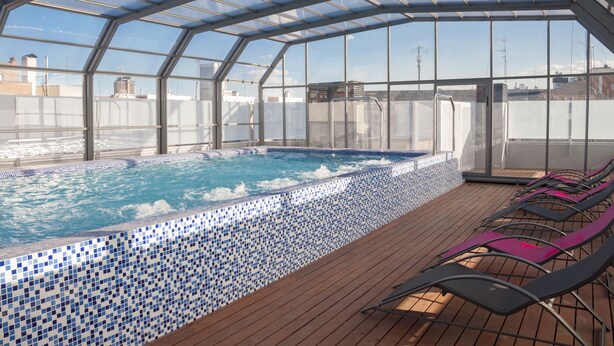 A rooftop swimming pool with a glass enclosure, wooden deck, and lounge chairs. - Funway Academic Resort - Adults Only (Madrid)