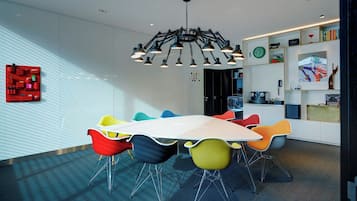Een moderne vergaderruimte met een grote tafel, kleurrijke stoelen, een bijzondere kroonluchter en een boekenplank vol diverse objecten.