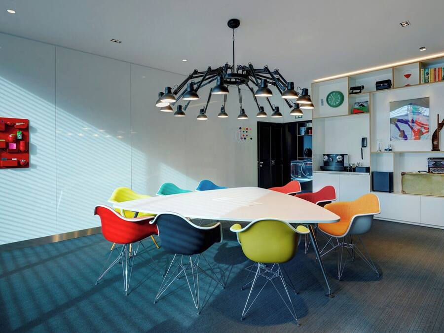 Een moderne vergaderruimte met een grote tafel, kleurrijke stoelen, een bijzondere kroonluchter en een boekenplank vol diverse objecten.