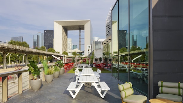 An outdoor seating area with tables and chairs, surrounded by large windows offering city views.