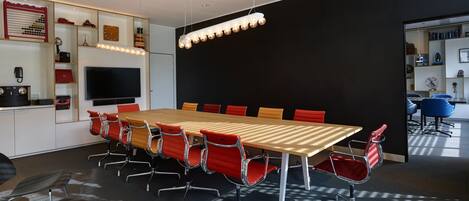 Une salle de conférence avec une longue table, des chaises rouges et noires, un téléviseur et des étagères remplies de divers objets.