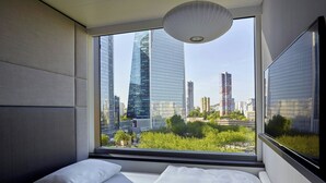 A modern hotel room with a large window offering a city view, a bed with white linens, and a hanging light fixture.