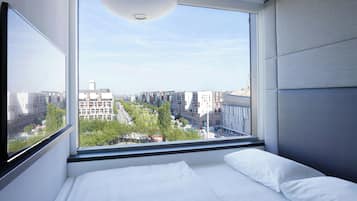 A modern hotel room with a large window offering a city view, a bed with white linens, and a ceiling light fixture.