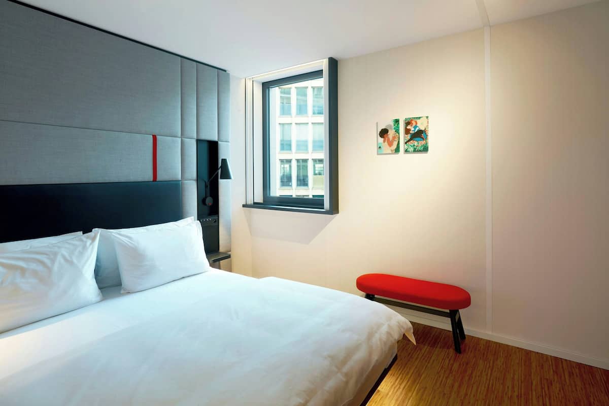 Et moderne hotelværelse med en stor seng, en rød bænk, et vindue med udsigt og kunst på væggen.