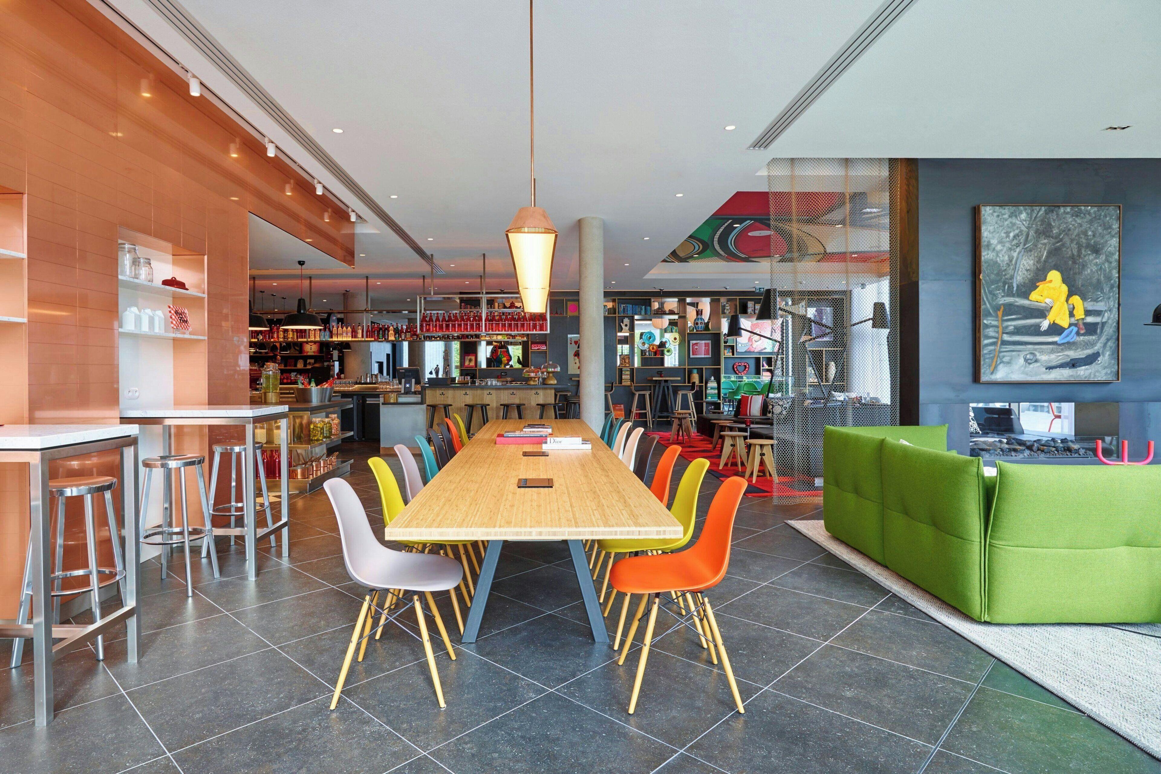 Un espace repas moderne avec une longue table en bois entourée de chaises colorées, un coin bar avec des tabourets et un coin salon avec des sièges rembourrés de couleur verte.