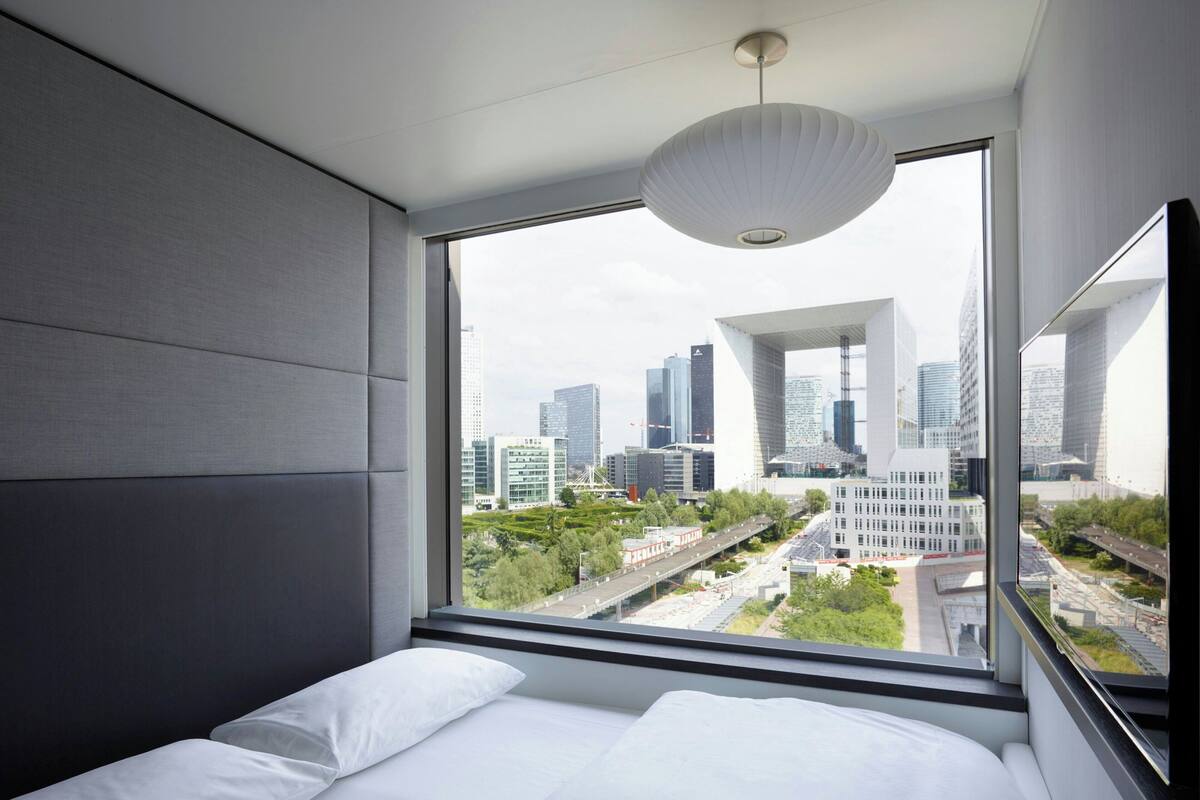 Habitación de hotel moderna con un amplio ventanal que ofrece vistas a la ciudad, una cama con ropa blanca y una lámpara de techo original.