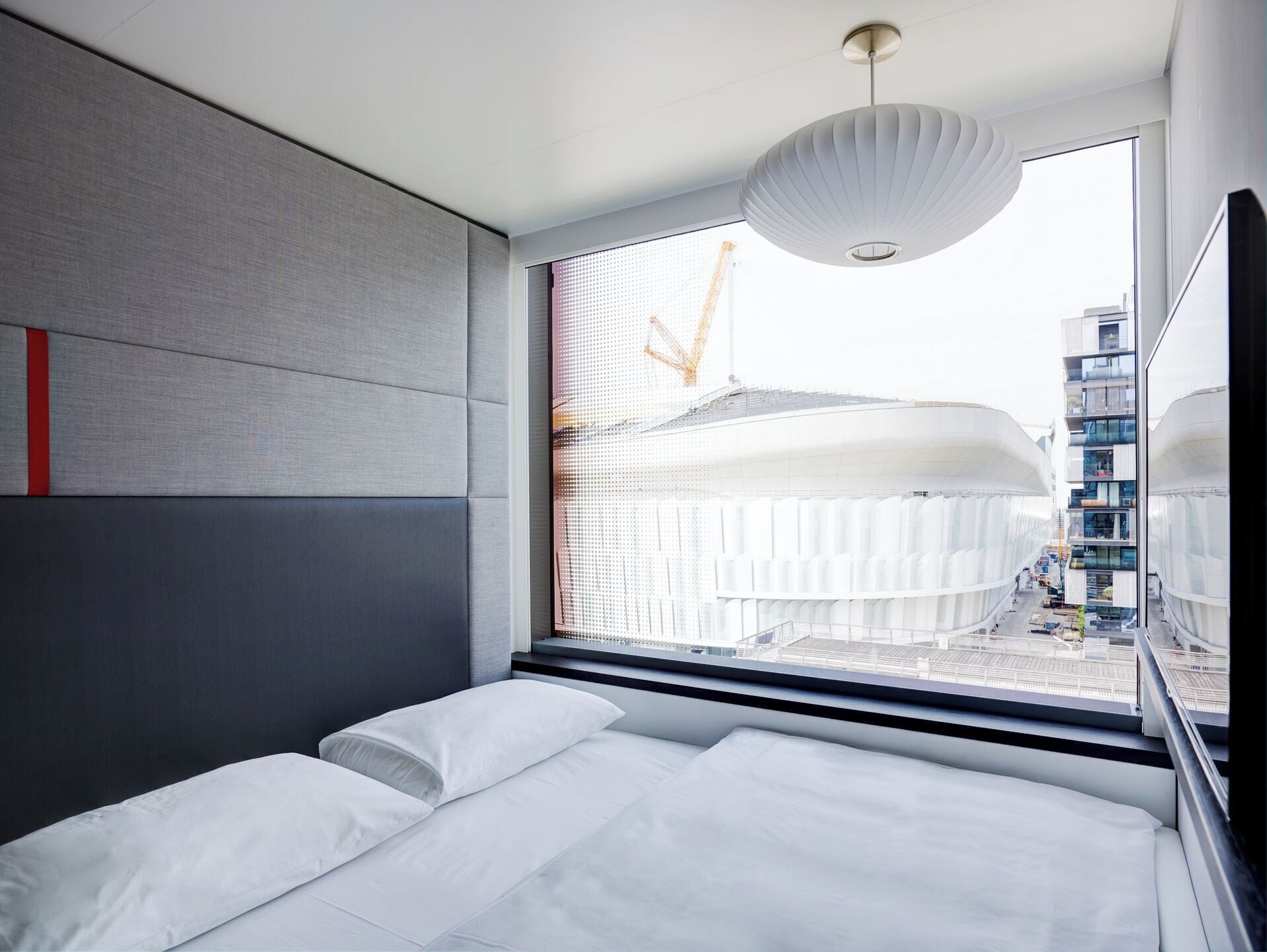 A modern hotel room with a large bed, a view of a construction site, and a unique ceiling light fixture.