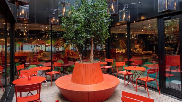 Un espace extérieur moderne, aménagé avec des tables et des chaises rouges, entouré de murs de verre et agrémenté d’un grand bac à plantes placé au centre.