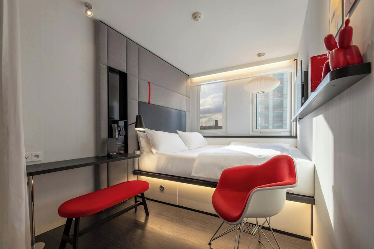 Quarto de hotel compacto com cama, escrivaninha, cadeira e janela com vista para a cidade.