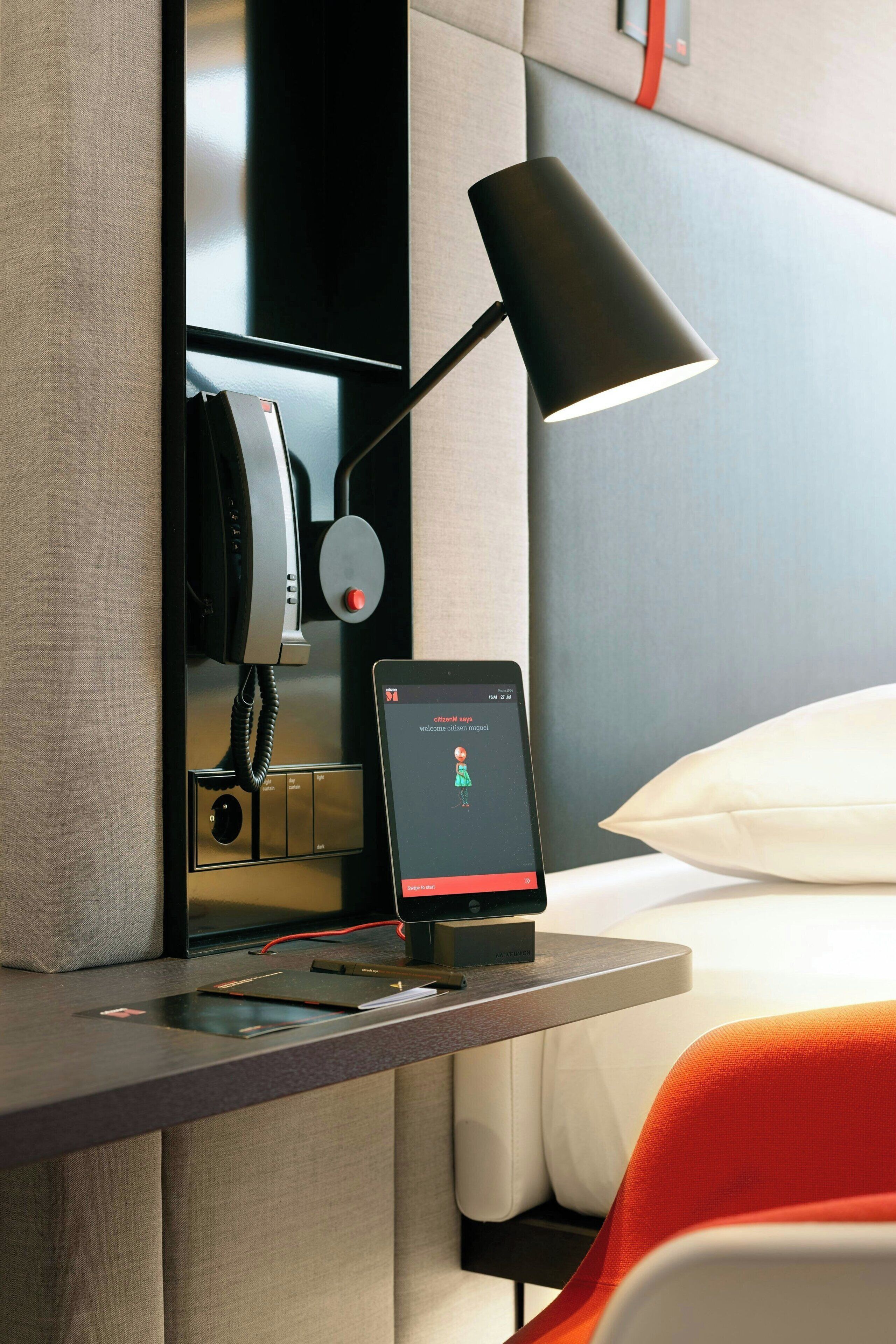 Una camera d'albergo con una scrivania, un telefono, un tablet e un letto con cuscini.