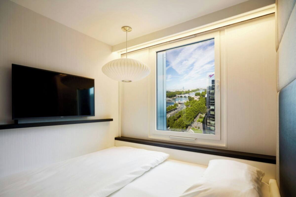 Quarto de hotel moderno com cama, TV de tela plana na parede, uma janela grande com vista para a cidade e uma luminária pendente.