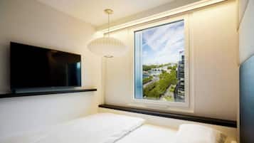 A modern hotel room with a bed, a flat-screen TV mounted on the wall, a large window with a cityscape view, and a pendant light.