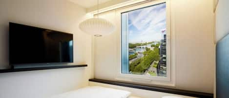 A modern hotel room with a bed, a flat-screen TV mounted on the wall, a large window with a cityscape view, and a pendant light.