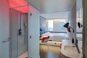 A modern hotel room with a glass shower enclosure, a bed with white bedding, a red chair, a large window, and a bathroom with a sink and mirror. - citizenM Paris Gare de Lyon (Paris)
