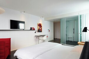 Una habitación de hotel moderna con una cama grande, un escritorio, una silla, un televisor y una zona de ducha con paredes de vidrio.