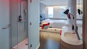 Une chambre d’hôtel moderne dotée d’une douche en verre, d’un lit avec une literie blanche, d’un fauteuil rouge et d’une grande fenêtre donnant sur la ville.