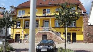A yellow building with a staircase leading to a balcony, a black car parked in front, and a sign for 'Hotel Zum Schwarzen'.
