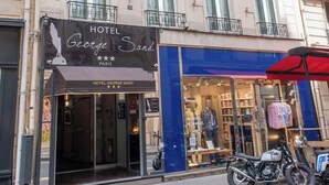 En hotels facade med et skilt, der viser "HOTEL GEORGE SAND PARIS", et butiksvindue med mannequiner, og en motorcykel parkeret udenfor.